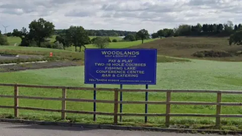 Google A blue sign saying Woodlands Golf and Country Club, above a fence with green grass and trees behind.