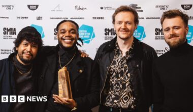 Four men are posing in front of a backdrop covered with logos and branding related to the Scottish music industry and the "Scottish Album of the Year Award." One of the men in the center is holding a wooden award.