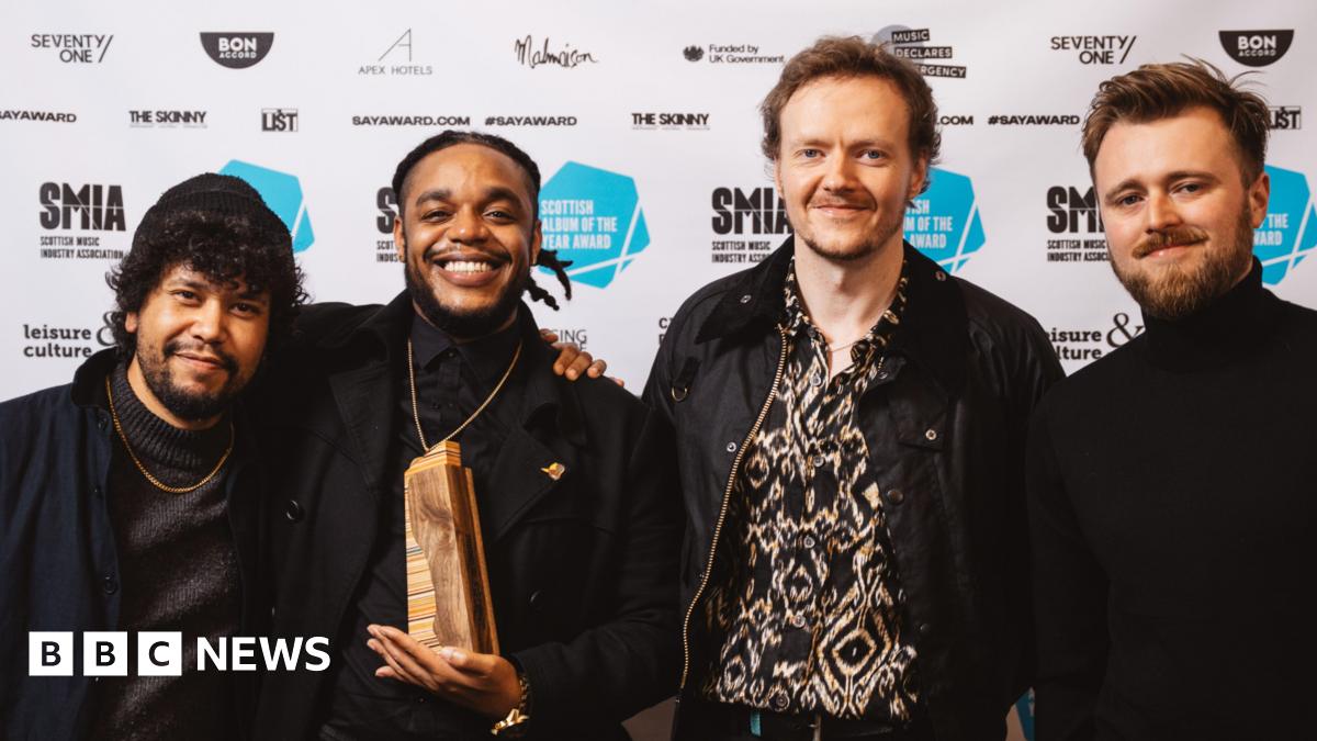 Four men are posing in front of a backdrop covered with logos and branding related to the Scottish music industry and the "Scottish Album of the Year Award." One of the men in the center is holding a wooden award.