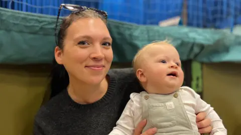 BBC Leigh Walker and her baby Clyde - A woman with sunglasses on her head smiling at the camera. She's black hair in a ponytail and a black jumper. She is holding on her lap her baby who is wearing green dungarees and a white baby-gro. He's looking away from the camera and has a tuft of blonde hair on his wee head. 