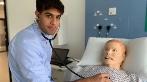 BBC A young man with dark hair and a blue shirt puts a stethoscope to the chest of a model of a patient used to train young doctors in a hospital bed.