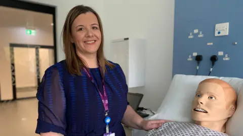 A woman in a blue dress with a purple lanyard round her neck with long, brown hair stands next to a model of 