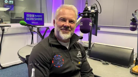 Ollie Phillips in the BBC Radio Berkshire studio, is smiling at the camera and wearing a black zipped jumper.