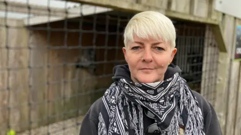 A woman stands in front of a wooden enclosure at a Bird of Prey visitor attraction. Nets form the walls of the enclosure, which contains two Great Grey Owls. One owl is just visible in the background, sitting on a perch. The woman is wearing a patterned black and white scarf.  