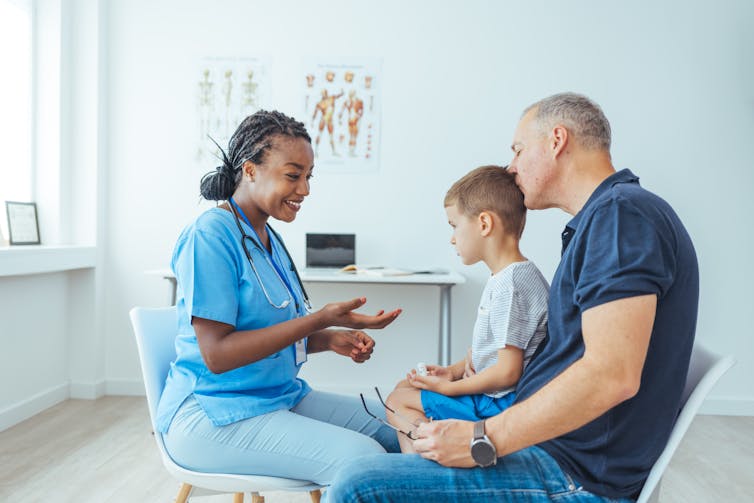 As a father comforts his young son with a kiss, a young female pediatrician smiles as she speaks to her young patient within a hospital setting.