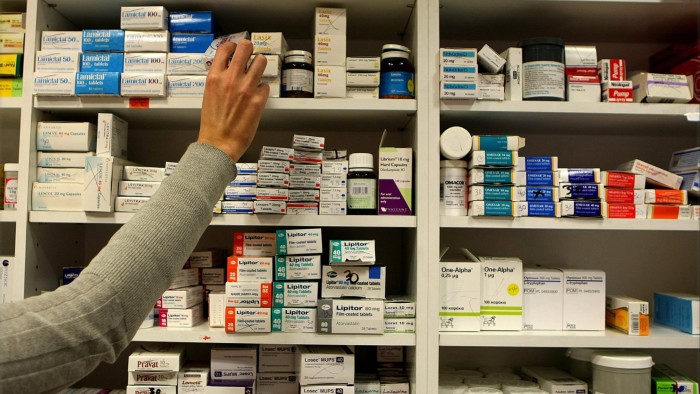 A pharmacist’s hand reaches for a medication box on shelves stocked with various prescription drugs.
