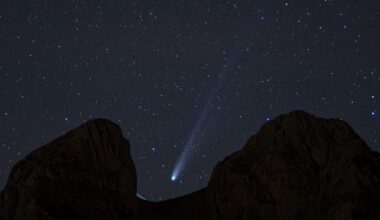 A white streak of a comet's tail can be seen in the center of the image as it streaks diagonally from top right to lower left between the dark silhouette of rocks blocking part of the starry night sky