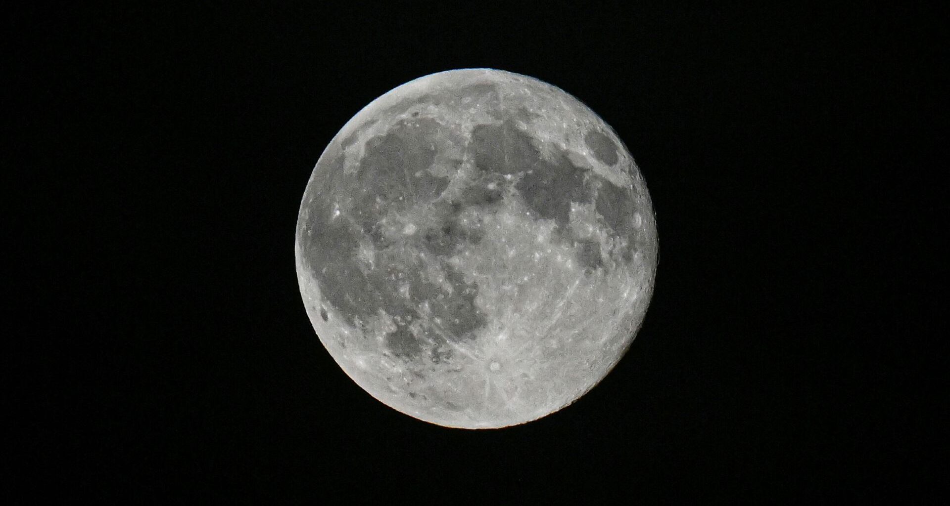 A detailed shot of a grey full moon is pictured against a black sky, sporting bright craters and dark features known as lunar maria.