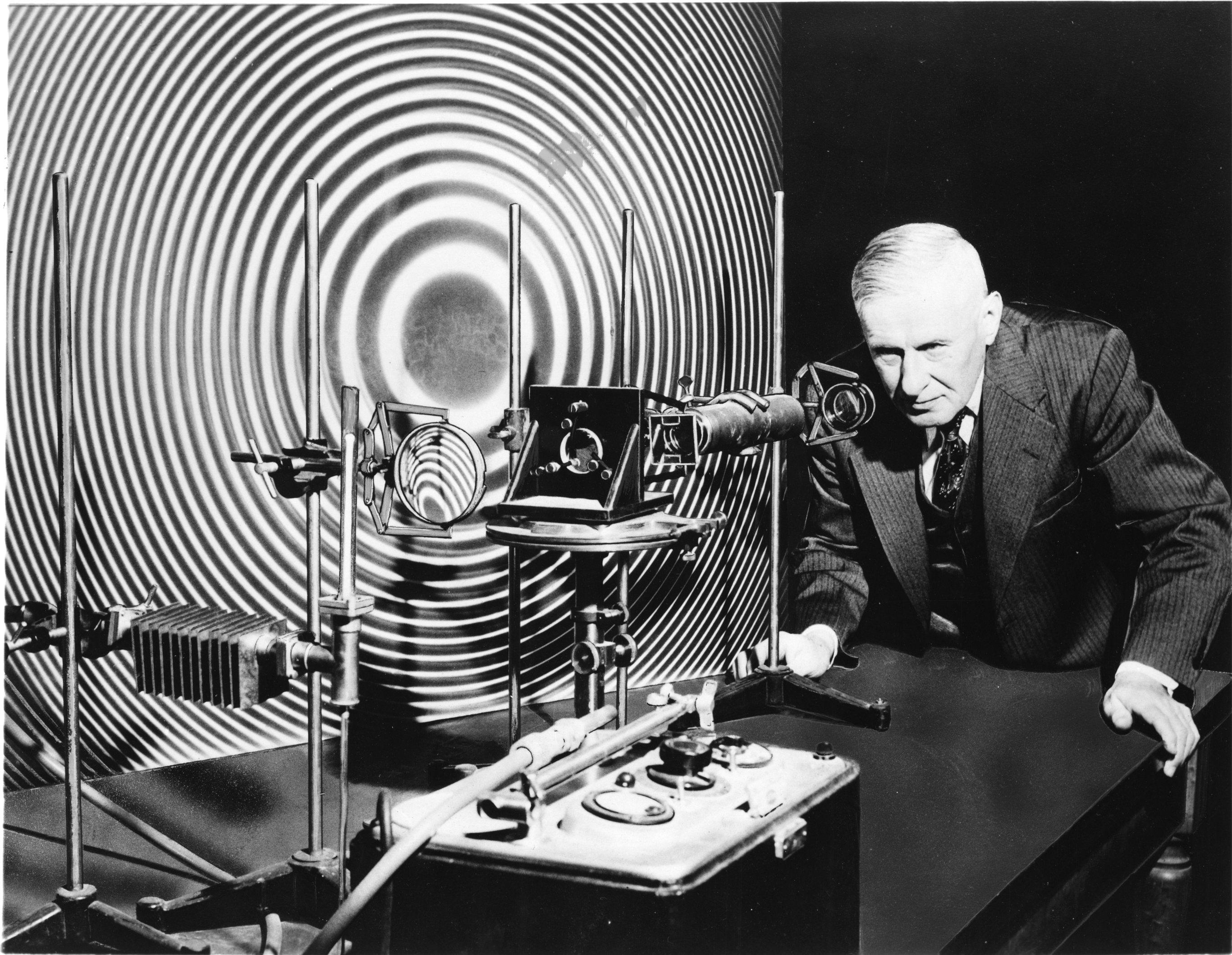 A man in a suit operates scientific equipment in front of a large circular interference pattern backdrop, using an instrument that appears to be one meter long, suggesting an experiment related to wave physics or optics.