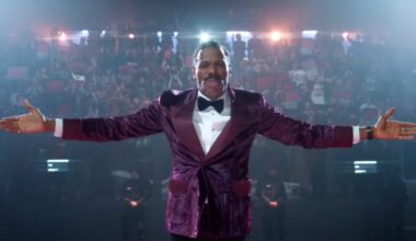 black man in colorful purple suit with arms outstretched in front of a camera with huge crowd behind him