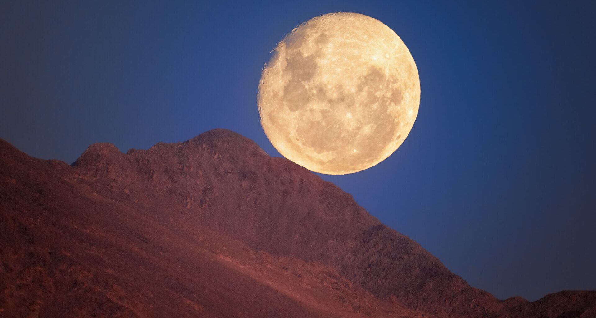 A glowing yellow moon is seen in a deep blue night sky with a red ridge of rock in front