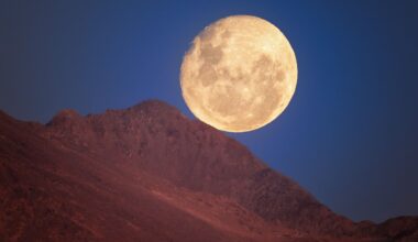 A glowing yellow moon is seen in a deep blue night sky with a red ridge of rock in front