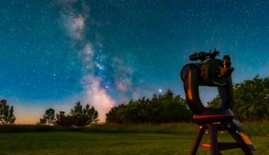 A telescope on a tripod sits in a grassy field looking at the pink glow of the Milky Way galaxy past some trees with a light blue night sky behind