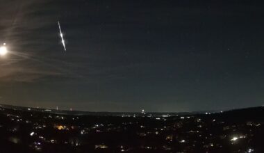 A bright fireball meteor is shown streaking earthward through the night sky over the lights of a town. The bright reflected light of the moon is visible to its left.