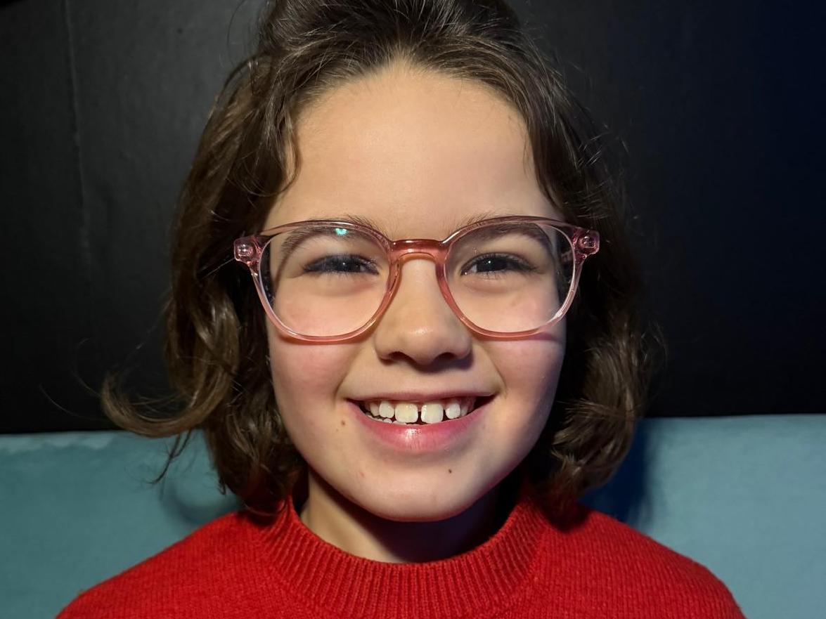Eden Dean smiling, wearing pink glasses and a red sweater with a reindeer design.