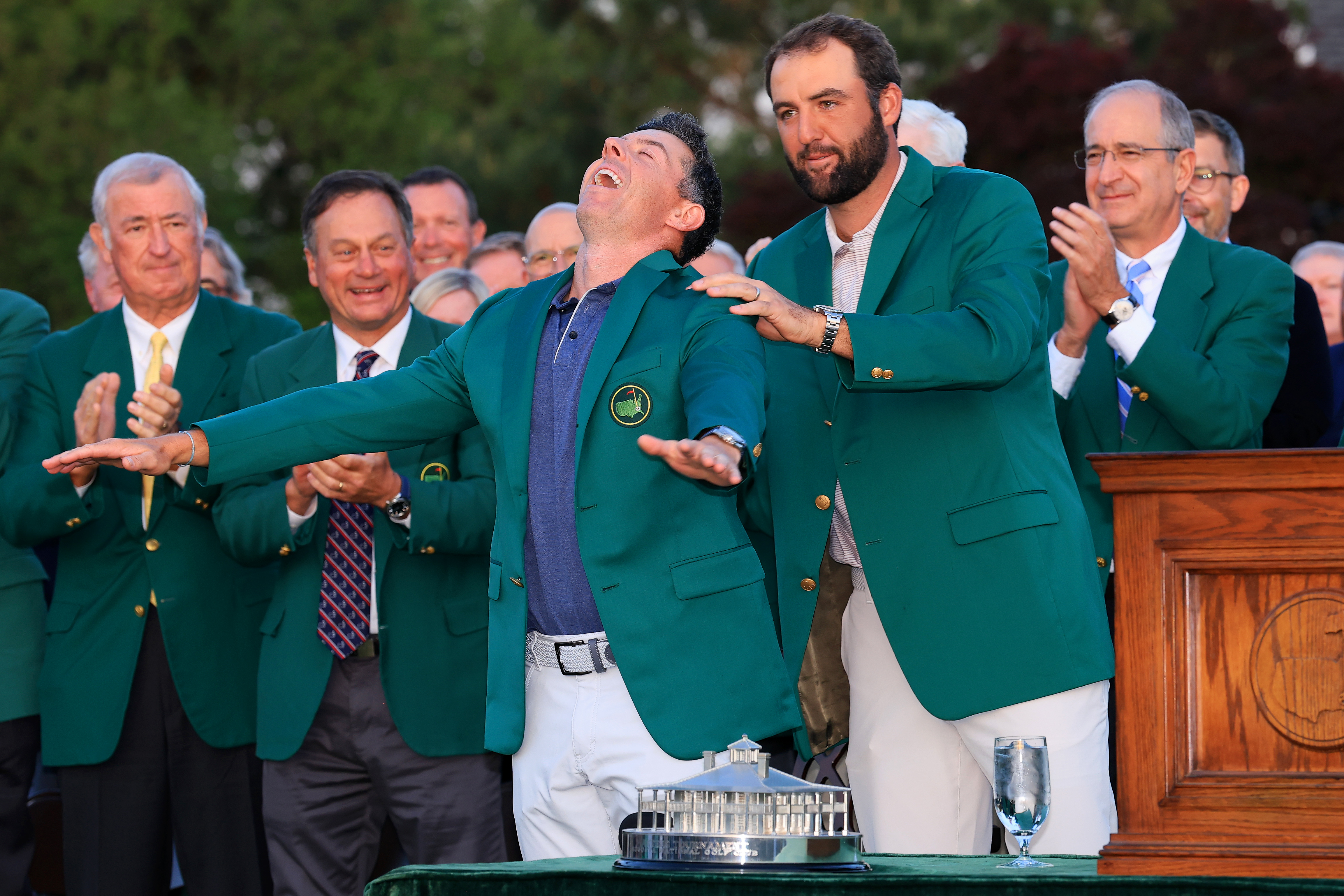 Rory McIlroy in a green jacket reacting joyfully as Scottie Scheffler helps him put it on.