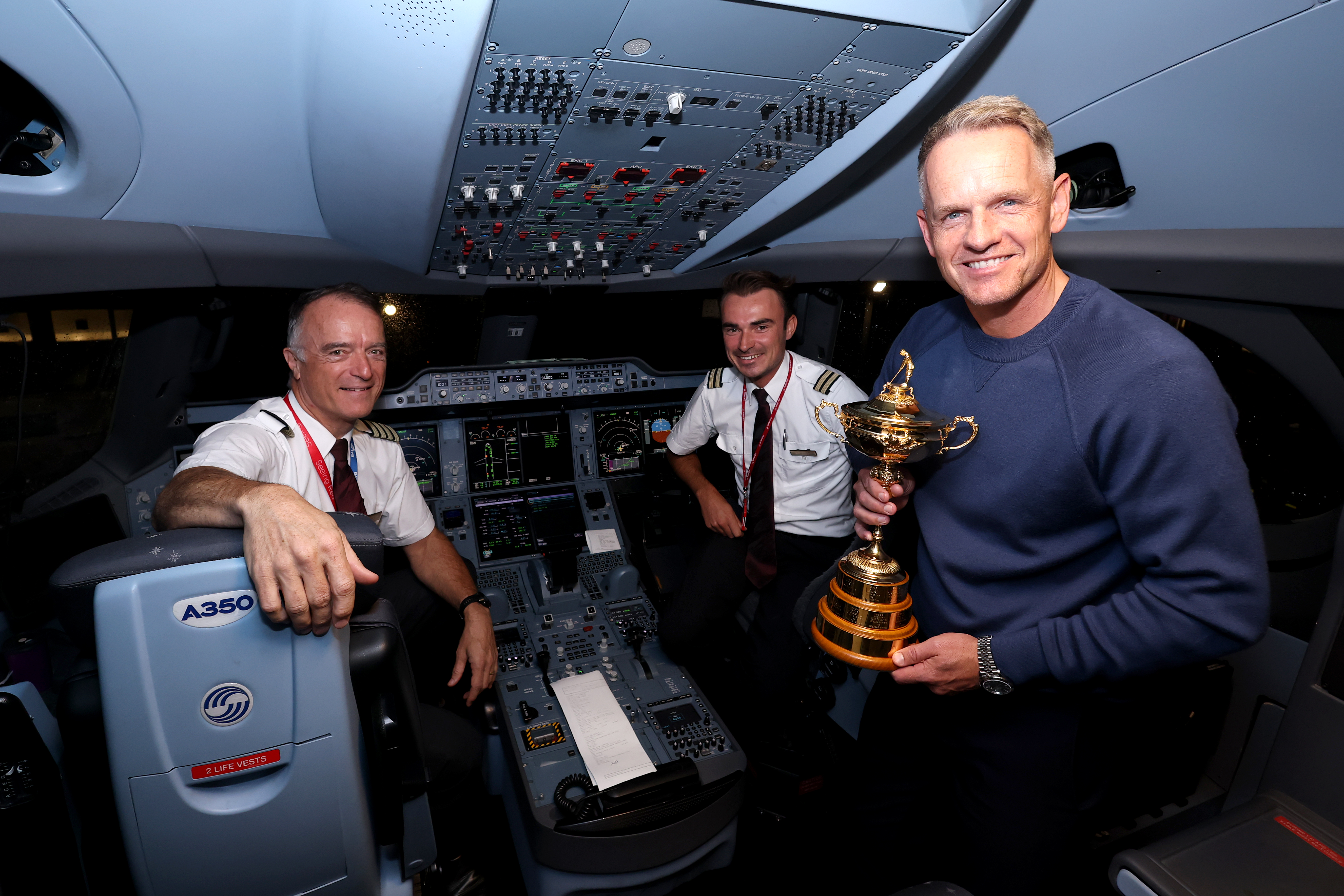 Luke Donald, Team Europe Captain, poses with the Ryder Cup trophy in an airplane cockpit with two pilots.