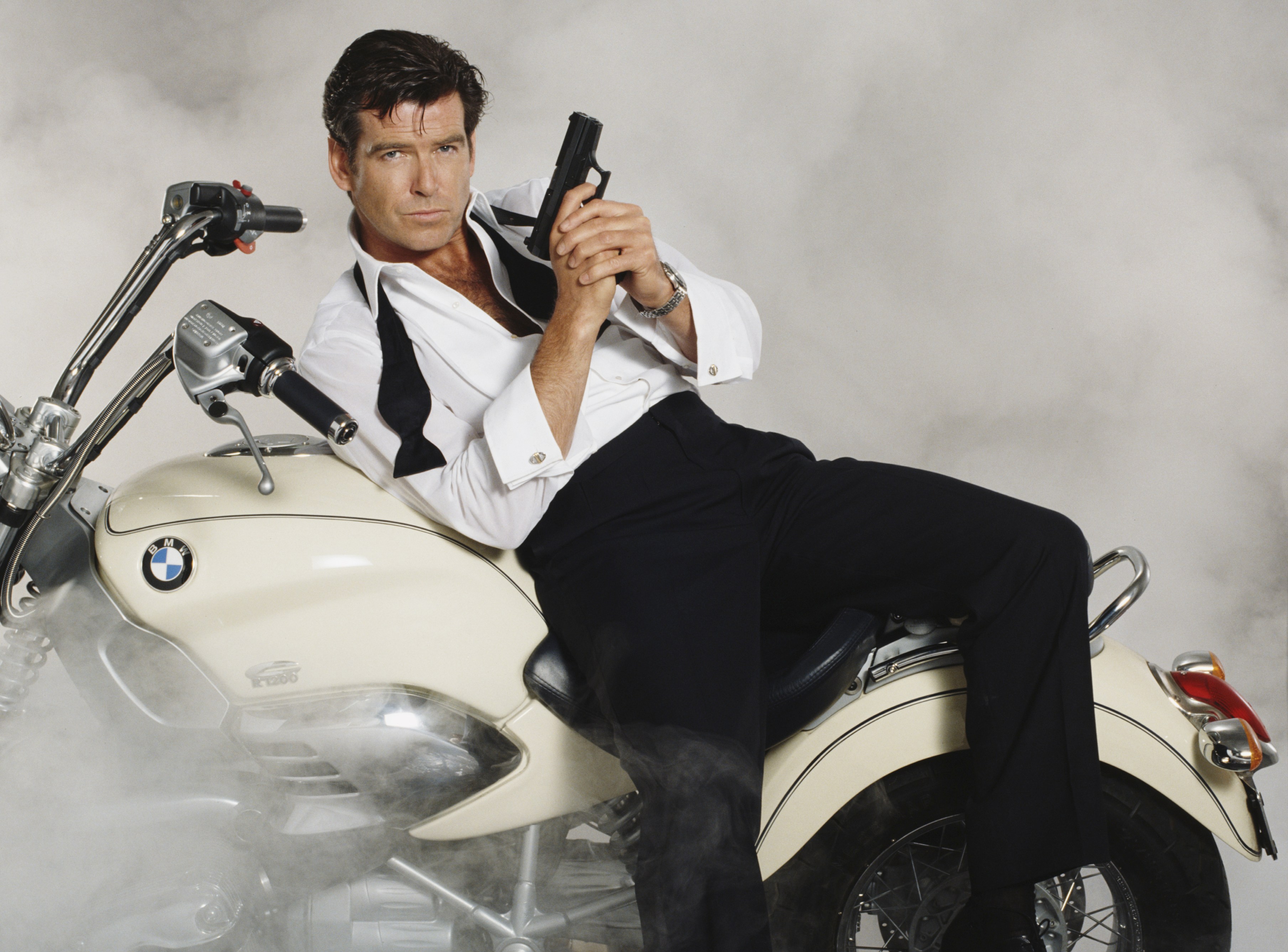 Pierce Brosnan as James Bond, in a white shirt and black trousers, holding a pistol and sitting on a white BMW motorcycle surrounded by smoke.