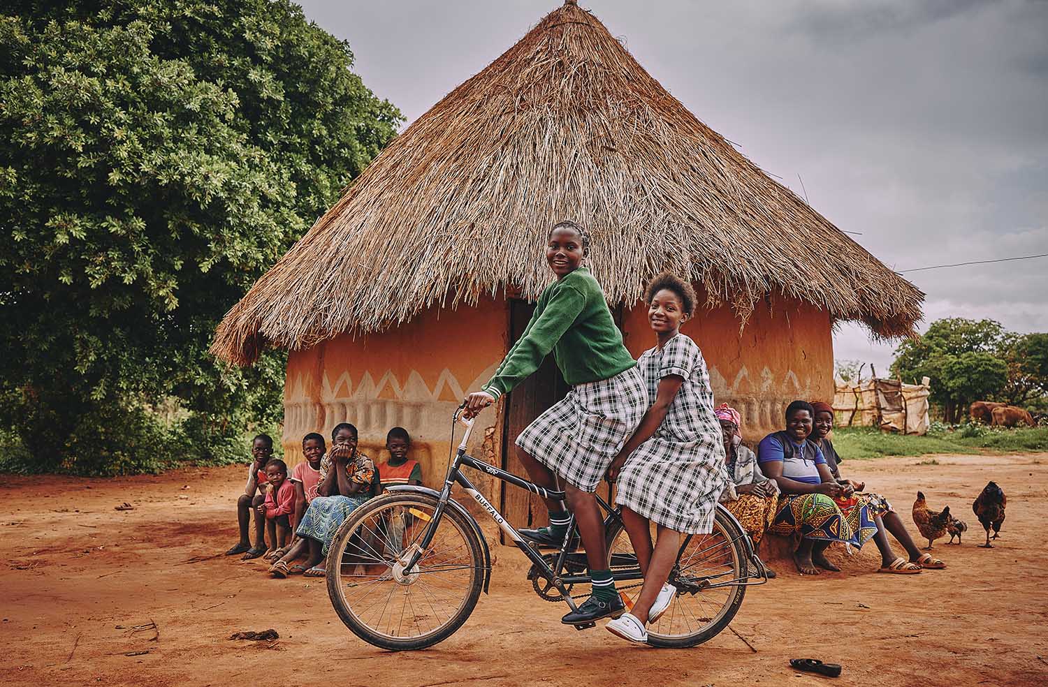 Schoolgirls Anastasia Mufwemba and Sylvia Komboni on a bicycle, with other children and adults watching, in front of a thatched-roof house.