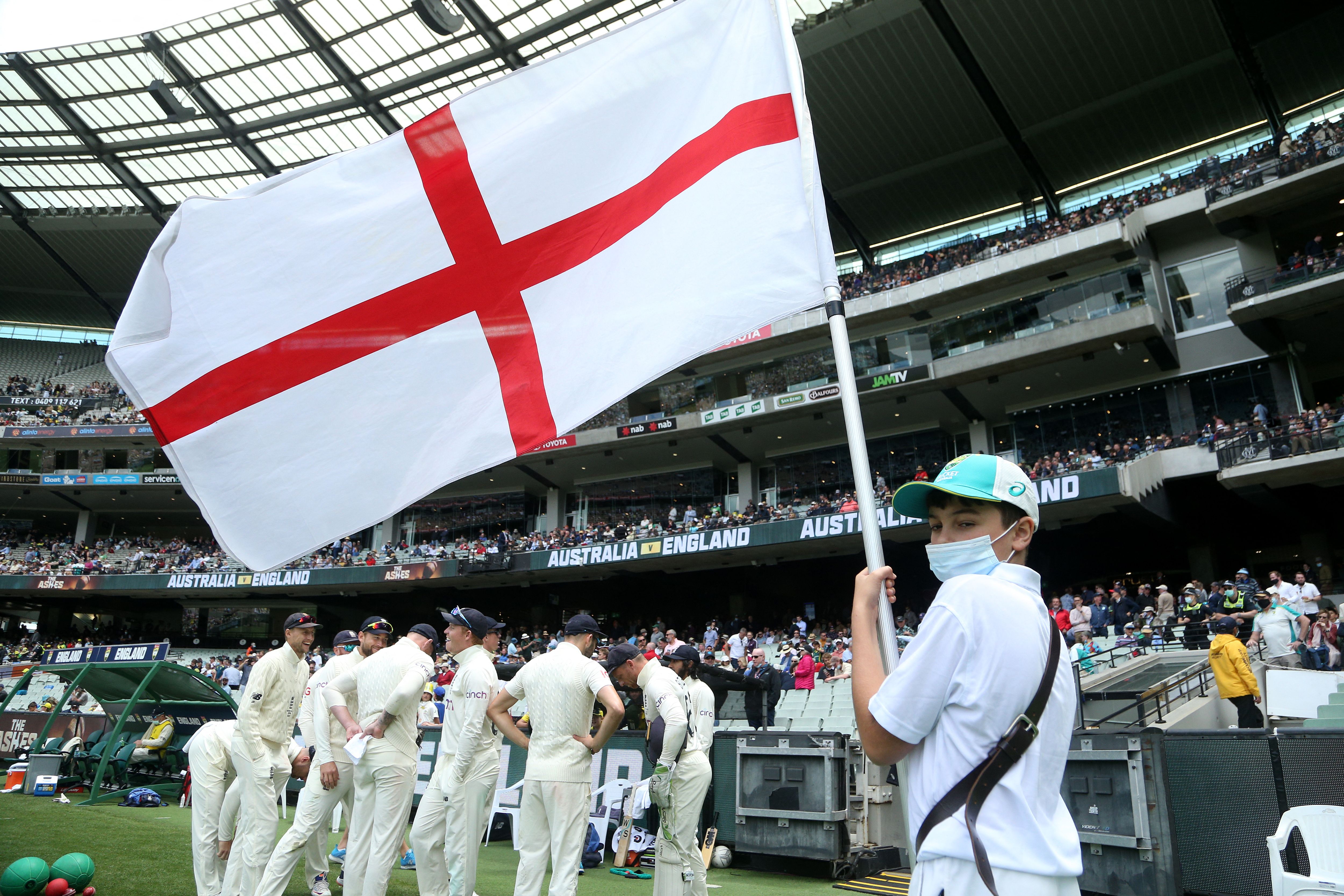 A masked flag bearer holds the England flag as the England cricket team stands on the field at the Melbourne Cricket Ground.