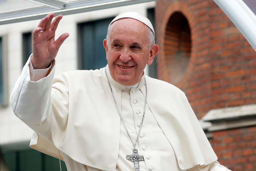 Pope Francis in white robes and wearing a whiet cap and silver cross chain waving with his right hand and smiling