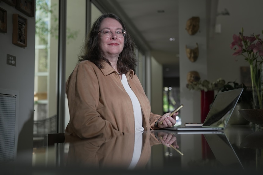 A woman with glasses and a brown jacket sits at a table with a laptop and mobile phone in front of her.