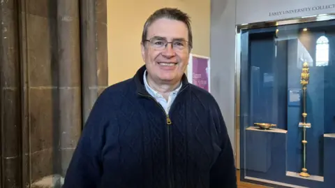 Dr Neil Clark, a man wearing a dark blue top and with short dark hair and glasses, standing in a museum. He is smiling into the camera.
