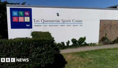 The outside of Les Quennevais Sports Centre. It has a blue box with different graphics inside of them representing a range of activities. The wall is white and in front of it is a bush and grass.