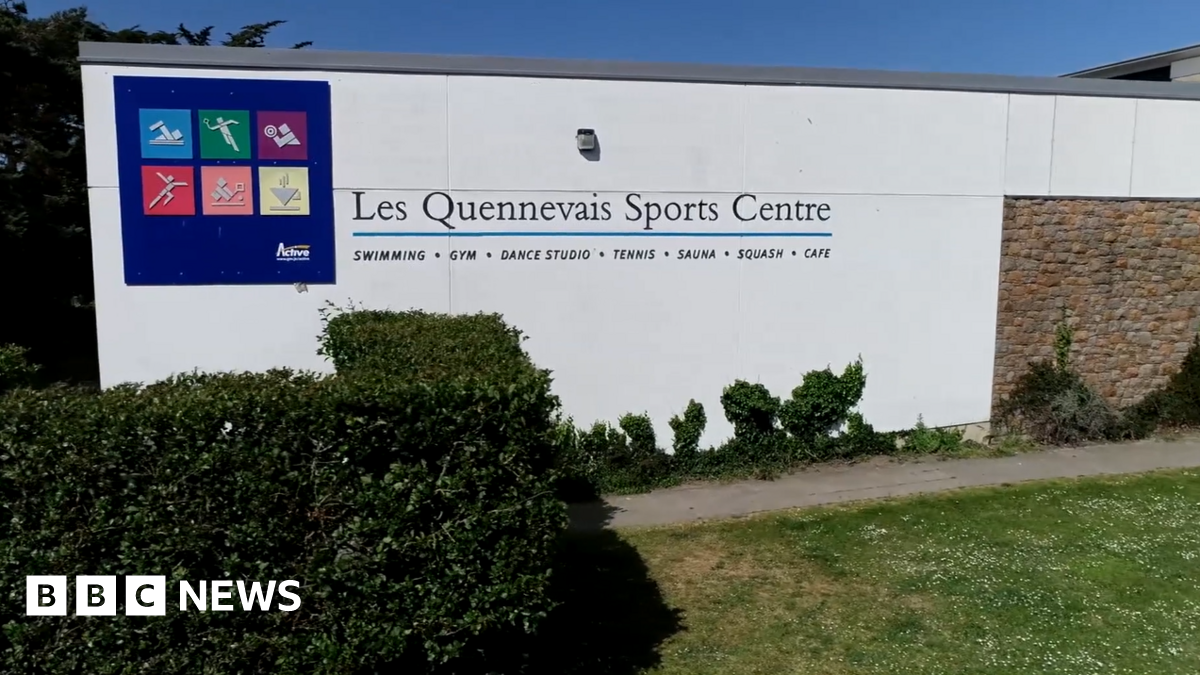 The outside of Les Quennevais Sports Centre. It has a blue box with different graphics inside of them representing a range of activities. The wall is white and in front of it is a bush and grass.
