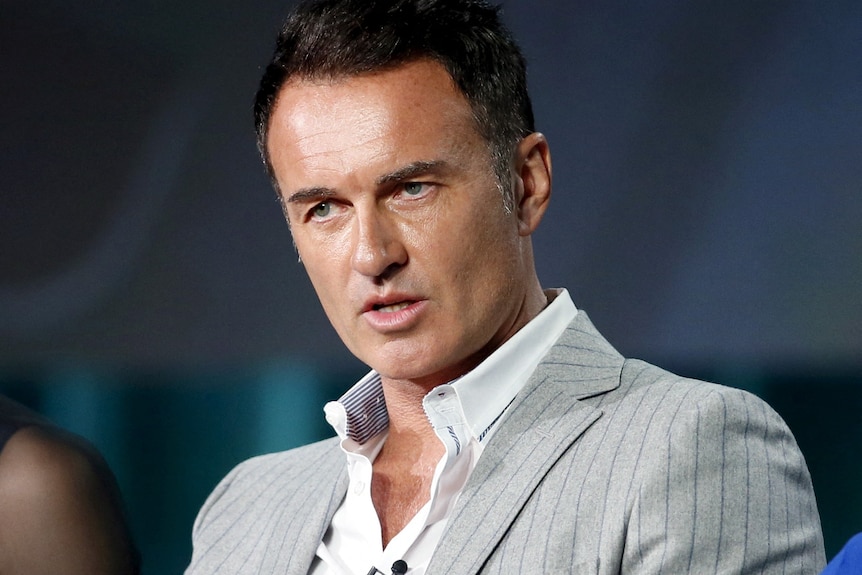 Julian McMahon speaking to someone while wearing a suit and a lapel microphone