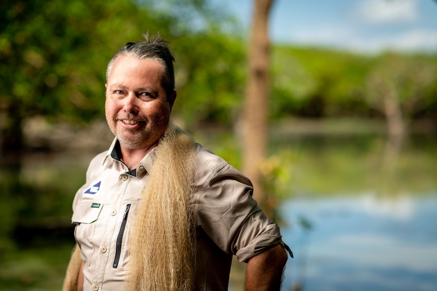 A portrait photo of a man smiling by a billabong. He wears a Charles Darwin University fishing shirt.