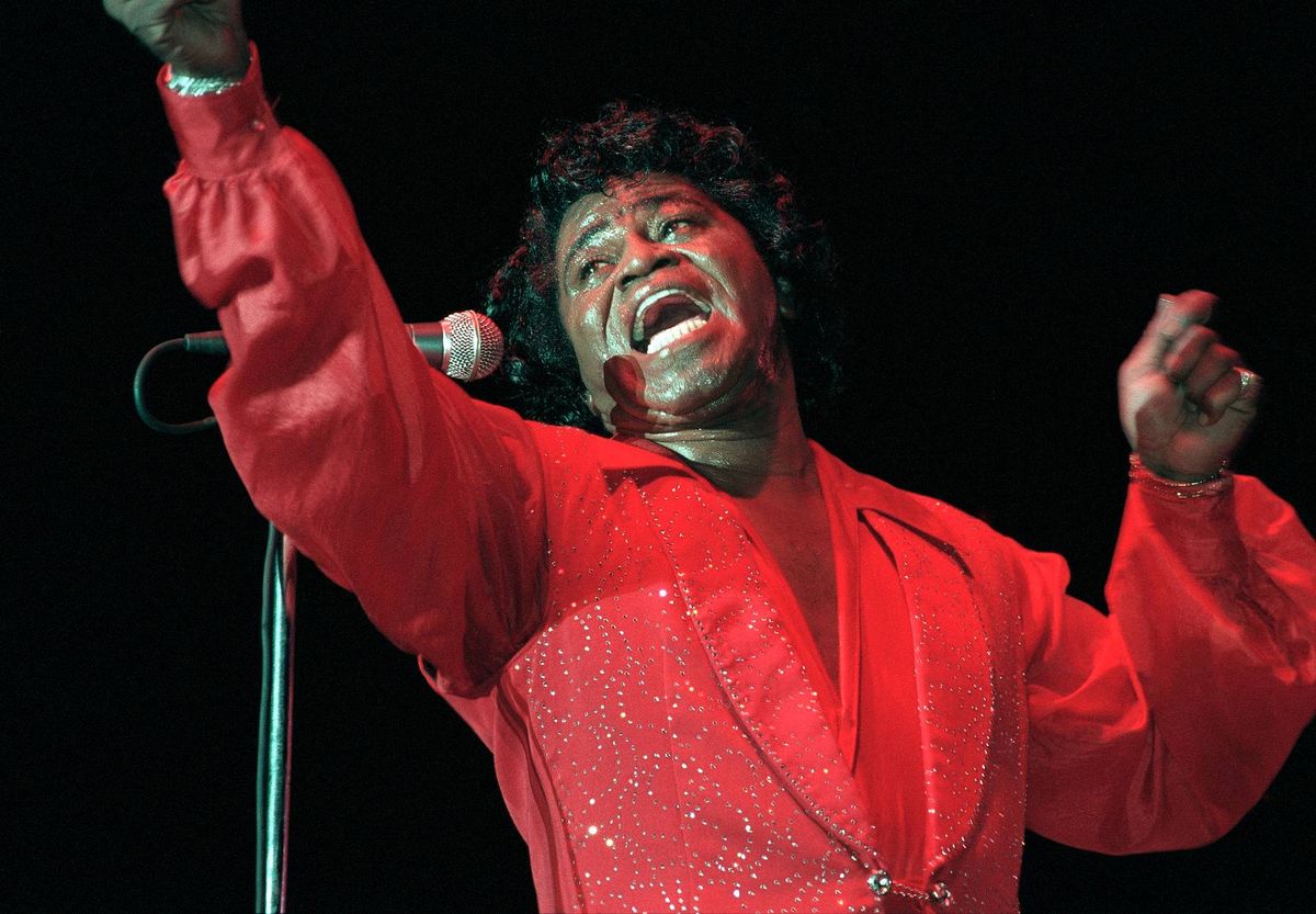 An individual is seen passionately performing on stage, clad in a vibrant red suit, with one arm raised and an open mouth, as they passionately engage with the audience. The setting is dimly lit, with a microphone positioned in front of them.