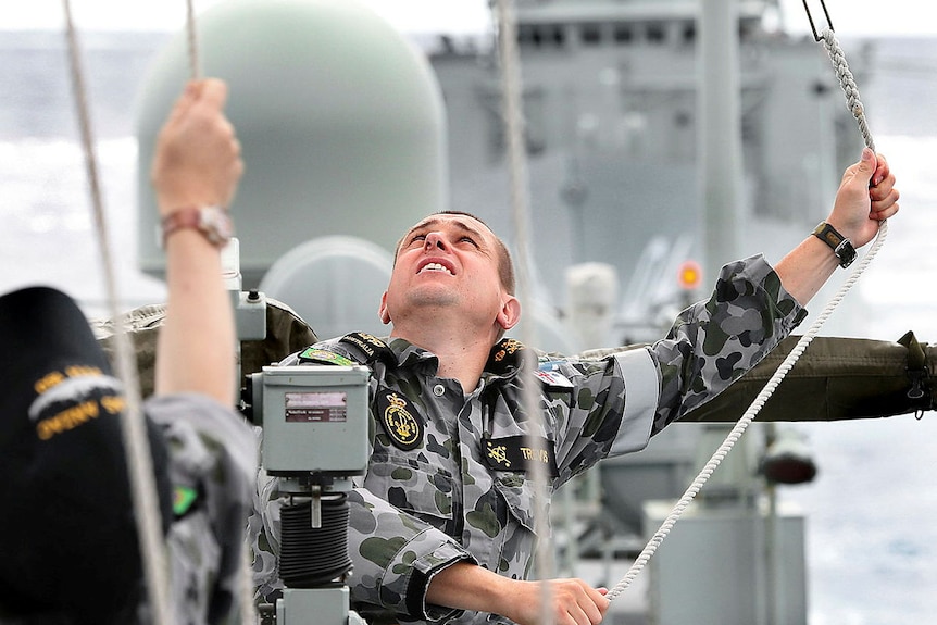 A man on a ship looking up as he holds and pulls ropes.