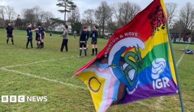 A number of players from the Brighton and Hove Sea Serpents rugby club on the pitch at Hove recreation ground. The players are all wearing navy kits and there is a club flag in the foreground of the image.