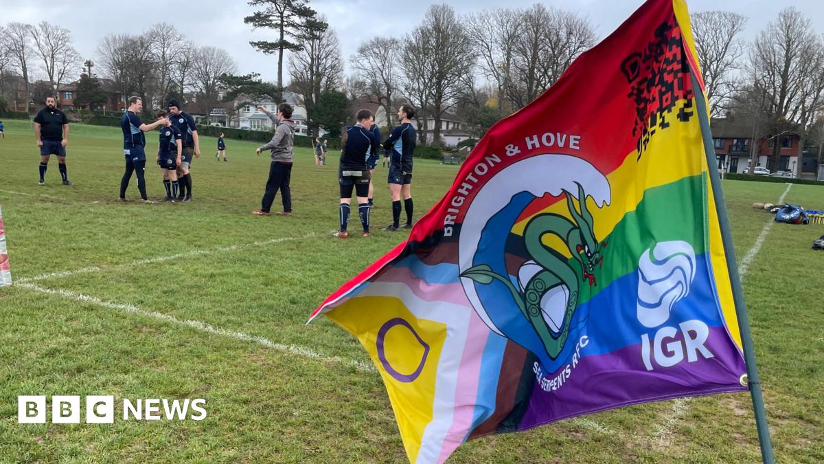 A number of players from the Brighton and Hove Sea Serpents rugby club on the pitch at Hove recreation ground. The players are all wearing navy kits and there is a club flag in the foreground of the image.