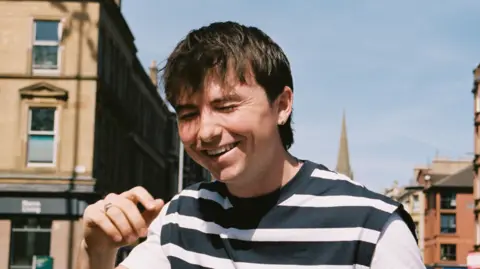 APB PR Shay O'Dowd, a young man with short dark hair and wearing a striped back and white top, laughing while standing outside 