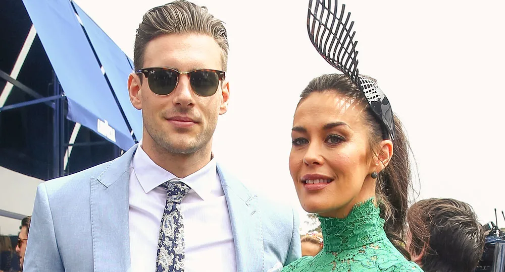 Two people at an outdoor event; man in a suit with sunglasses, woman in green dress with decorative headpiece.