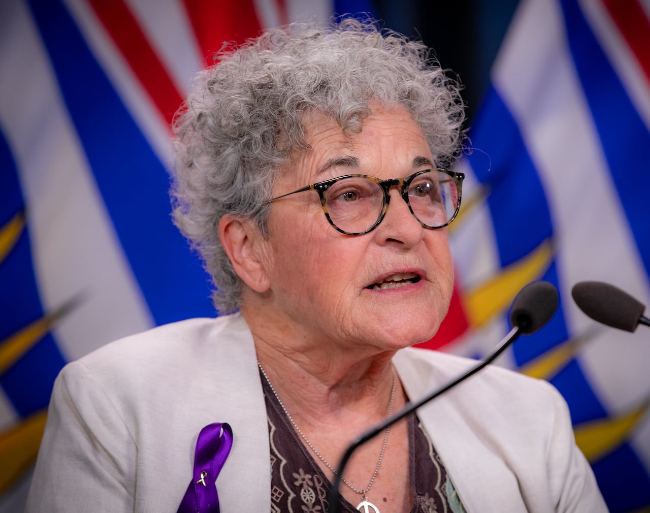 A white woman with short curly hair speaks in front of B.C. flags.