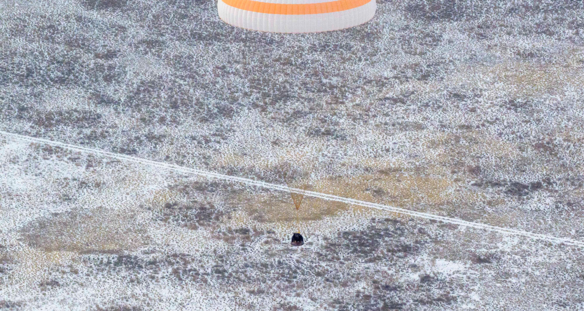An orange and white striped parachute attached to a black spacecraft. The spacecraft is very small compared to the parachute. It is landing on ground that has white frost on it.