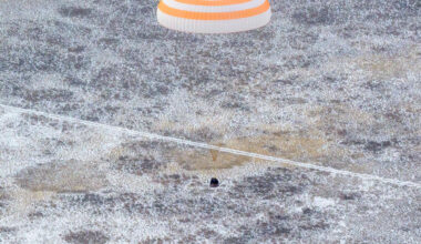 An orange and white striped parachute attached to a black spacecraft. The spacecraft is very small compared to the parachute. It is landing on ground that has white frost on it.