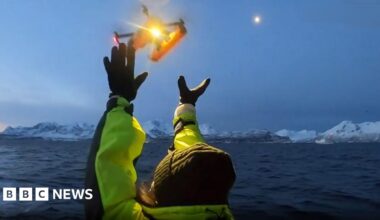 A research scientist dressed in a wet weather gear holds a drone on a boat. Behind is a choppy sea with ice flows.