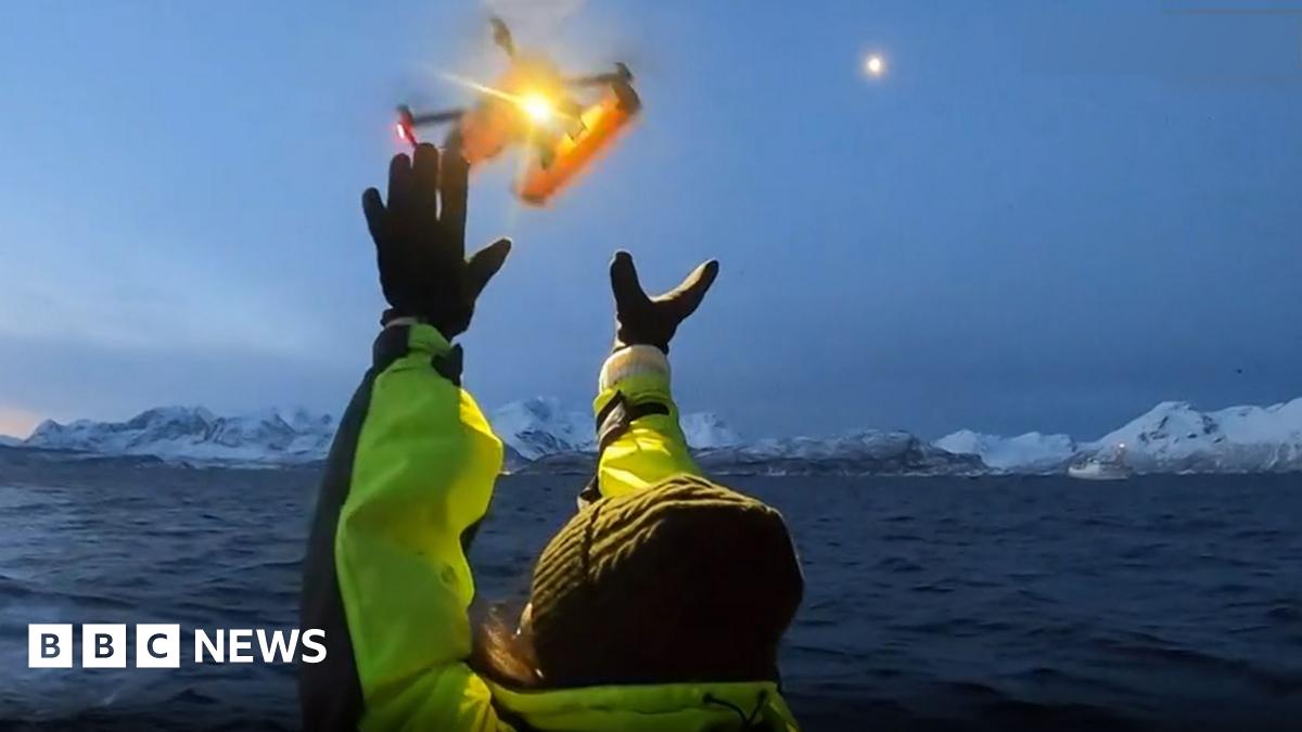 A research scientist dressed in a wet weather gear holds a drone on a boat. Behind is a choppy sea with ice flows.