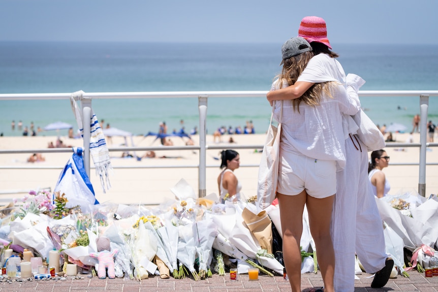 Two people comfort each other while looking at bouquets of flowers at Bondi Beach