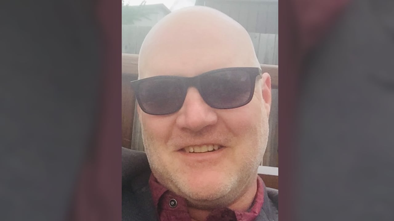 A man in a dark suit jacket and maroon-coloured collared shirt.