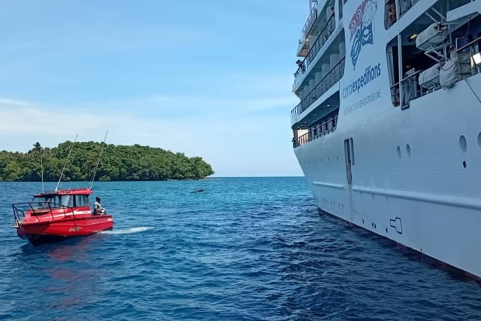 A small red boat alongside a large white cruise ship on water, with an island in the background.
