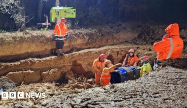 Four men in high-vis orange clothing stand next to a large pipe in an excavated trench. It is dark but the work site is brightly lit. Two other men are stood higher up the banks of the trench.