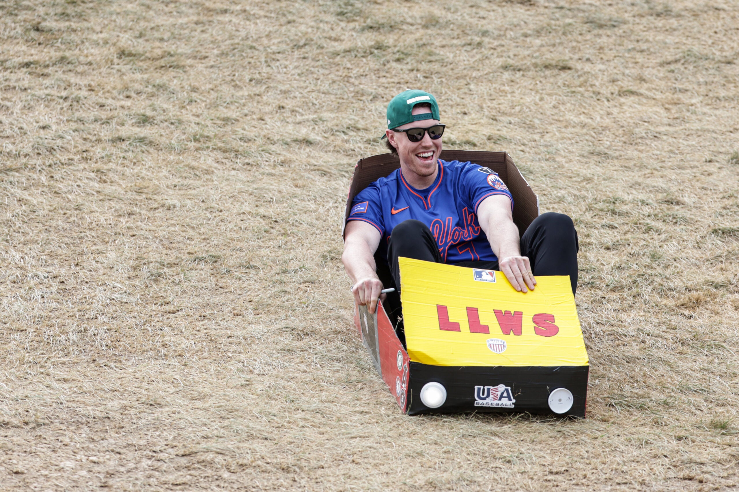 A man wearing sunglasses and a green cap slides down a grassy hill in a cardboard box decorated to look like a car with LLWS written on the front.