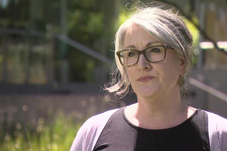 A woman with blonde hair, wearing glasses, a black top and pink cardigan stands outside on a sunny day.