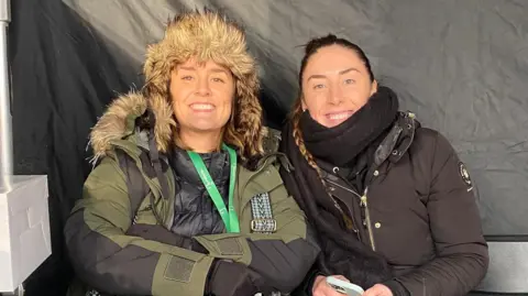Olivia Jerrard Alice Jones and Olivia Jerrard are sitting next to each other in a tent on set. They are both wrapped up warm in large coats, scarves and hats. They are both looking directly at the camera and are smiling.
