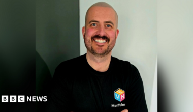 Jack Woodhams smiles at the camera. he has a shaved head and a short, dark beard and wears a black t-shirt with the Menfulness logo on the chest - a cube with blue, orange and pink sides and white lettering below spelling out Menfulness.